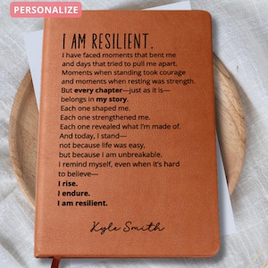 Puede incluir: Un diario de cuero marr&oacute;n con las palabras "I AM RESILIENT" impresas en la portada. El diario contiene un poema sobre la resiliencia y est&aacute; personalizado con el nombre "Kyle Smith". Una cinta roja como marcap&aacute;ginas es visible.