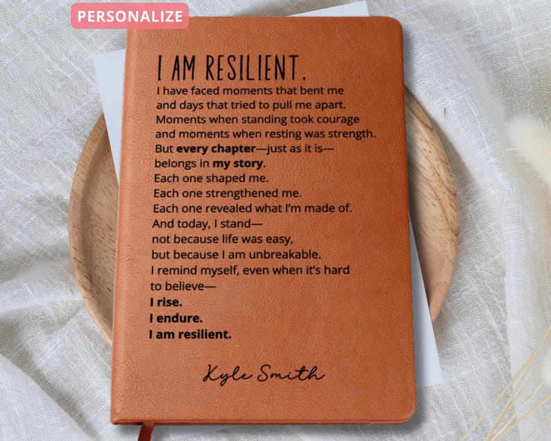 Puede incluir: Un diario de cuero marr&oacute;n con las palabras "I AM RESILIENT" impresas en la portada. El diario contiene un poema sobre la resiliencia y est&aacute; personalizado con el nombre "Kyle Smith". Una cinta roja como marcap&aacute;ginas es visible.