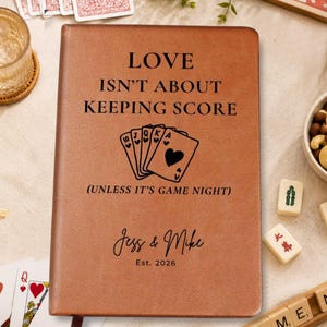 May include: A tan leather-like journal with black text that reads LOVE ISN'T ABOUT KEEPING SCORE (UNLESS IT'S GAME NIGHT) above an illustration of five playing cards, with the names Jess & Mike and the text Est. 2026 printed below.