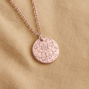 May include: Rose gold-colored pendant necklace featuring a circular charm with a detailed constellation design. The chain and pendant have a shiny finish. The necklace is displayed on a tan fabric surface.