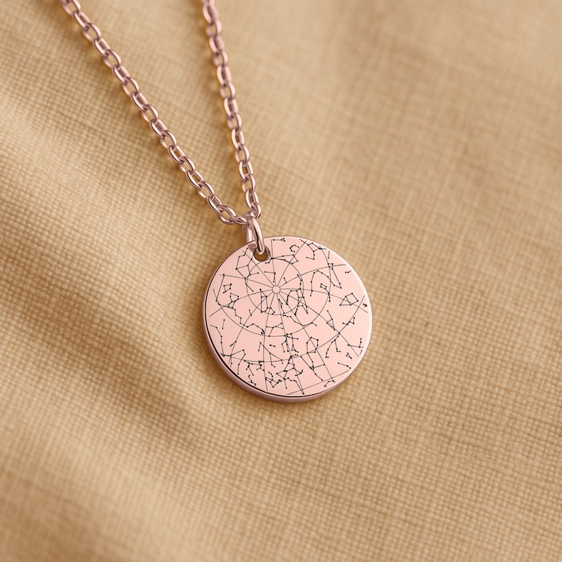 May include: Rose gold-colored pendant necklace featuring a circular charm with a detailed constellation design. The chain and pendant have a shiny finish. The necklace is displayed on a tan fabric surface.