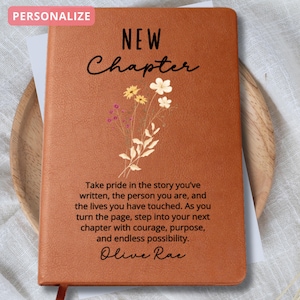 May include: A brown leather-bound journal with the words "NEW Chapter" and floral illustrations. The journal includes inspirational text and the word "PERSONALIZE" in a pink box. A red ribbon bookmark is visible.