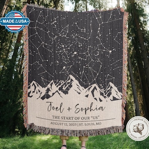 May include: A dark gray woven throw blanket featuring a constellation design with white stars and mountain silhouettes. The bottom section is beige with the names "Joel + Sophia" and the text "THE START OF OUR US" with a date and location.