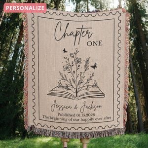 May include: Beige woven throw blanket with a black floral and book design. The blanket features the text "Chapter One", names, and a date. The edges have a colorful fringe.