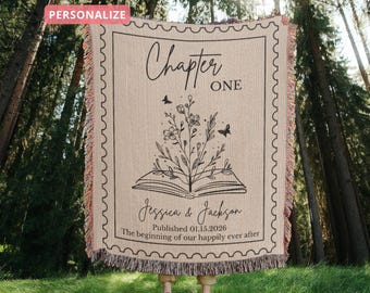 May include: Beige woven throw blanket with a black floral and book design. The blanket features the text "Chapter One", names, and a date. The edges have a colorful fringe.