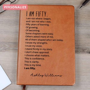 May include: A brown leather journal with the text "I AM FIFTY" and a poem about life's journey. The journal is personalized with the name "Ashley Williams". A pink rectangle with the word "PERSONALIZE" is in the top left corner.