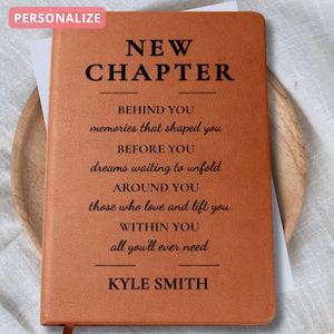 May include: A personalized brown leather journal with the words "NEW CHAPTER" at the top. Below, inspirational text is printed, followed by a name. The journal is open, revealing white pages, and rests on a wooden plate.