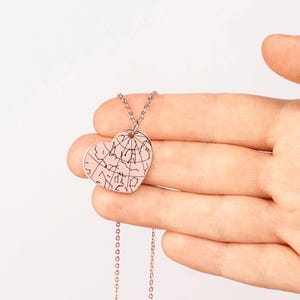 Puede incluir: Un collar con colgante en forma de coraz&oacute;n con un fondo rosa claro y un dise&ntilde;o de constelaci&oacute;n negro. El colgante cuelga de una fina cadena plateada. El collar se sostiene en una mano sobre un fondo blanco.