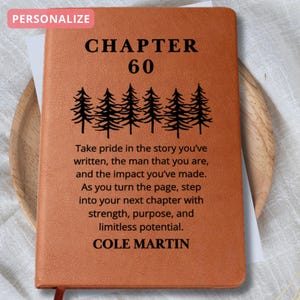 May include: A personalized, brown leather journal with the text "CHAPTER 60" and a row of pine trees. The journal includes inspirational text and the name "COLE MARTIN". A red ribbon bookmark is visible.