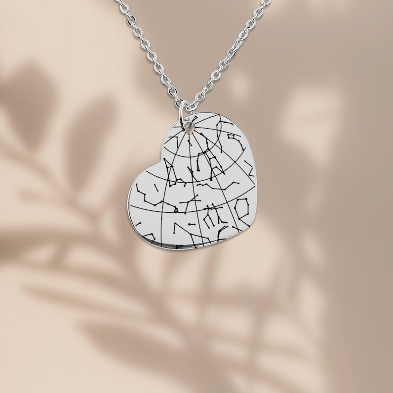 Puede incluir: Un collar plateado con un colgante en forma de coraz&oacute;n. El colgante presenta un dise&ntilde;o de constelaci&oacute;n en l&iacute;neas negras sobre un fondo plateado. La cadena del collar tambi&eacute;n es plateada y cuelga de la parte superior del colgante.
