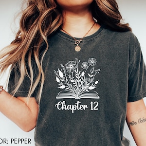 May include: Pepper-colored t-shirt featuring a white floral design emerging from an open book, with the text "Chapter 12" below. The shirt is a dark gray color and has a casual, relaxed fit.