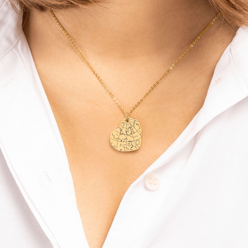 Puede incluir: Un collar dorado con un colgante en forma de coraz&oacute;n. El colgante presenta un dise&ntilde;o de mapa con l&iacute;neas finas. El collar se lleva sobre una camisa blanca.