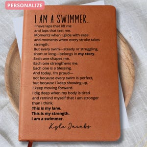 May include: A personalized brown leather journal with the text "I AM A SWIMMER" and a poem about swimming. The journal has a red ribbon bookmark and is displayed on a wooden plate. The word "PERSONALIZE" is in a pink box at the top.