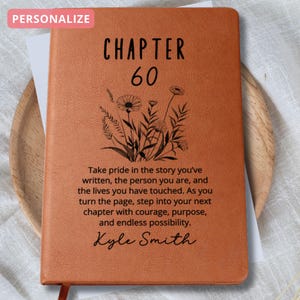 May include: A personalized, orange leather-bound journal with the text "CHAPTER 60" and a floral design. The journal includes a quote about pride and new chapters, with a name at the bottom. A red ribbon bookmark is visible.