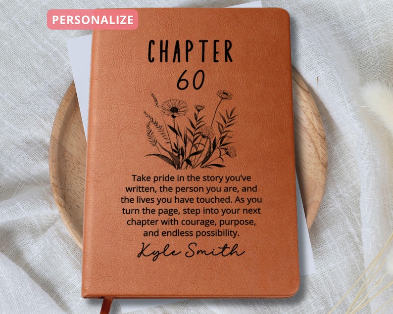 May include: A personalized, orange leather-bound journal with the text "CHAPTER 60" and a floral design. The journal includes a quote about pride and new chapters, with a name at the bottom. A red ribbon bookmark is visible.