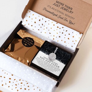 May include: An open gift box with a necklace and a gold-colored pouch. The necklace has a round pendant with a detailed design. The box has text that reads "MORE THAN JUST JEWELRY" and "Personalized Just For You!"