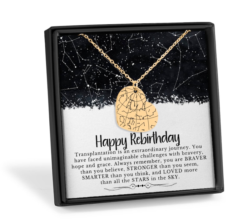 Puede incluir: Un collar con colgante dorado en forma de coraz&oacute;n con un dise&ntilde;o de constelaci&oacute;n, presentado en una caja de regalo negra. La caja contiene una tarjeta con el texto "Happy Rebirthday" y un mensaje inspirador sobre la valent&iacute;a.