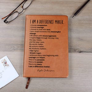 May include: A brown leather journal with the text "I AM A DIFFERENCE MAKER" and inspirational text. The journal has a ribbon bookmark and is next to a pen, glasses, and an envelope.