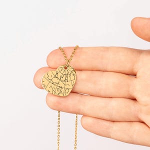 Puede incluir: Un collar dorado con un colgante en forma de coraz&oacute;n. El colgante presenta un dise&ntilde;o de mapa detallado. El collar se sostiene en una mano sobre un fondo blanco. La cadena es delicada y el colgante mide aproximadamente 3,8 cm de ancho.