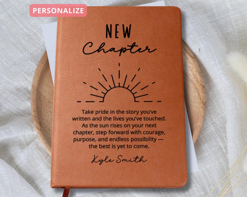 May include: A tan leather journal featuring the words "NEW Chapter" and a sunburst graphic. The journal has the text "Take pride in the story you've written...the best is yet to come." and the name "Kyle Smith".