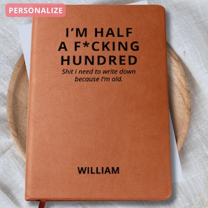 May include: A brown leather-bound notebook with the text "I'M HALF A F*CKING HUNDRED" and "Shit I need to write down because I'm old." The name "WILLIAM" is printed at the bottom. A red ribbon bookmark is visible.