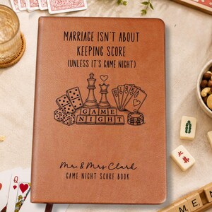 May include: A tan leather-bound score book featuring the text "Marriage isn't about keeping score (unless it's game night)" and "Mr. & Mrs. Clark Game Night Score Book" with engraved illustrations of chess pieces, playing cards, and dice.