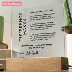 Könnte beinhalten: Transparente Acrylplatte mit den schwarzen Worten "DIFFERENCE MAKER". Die Platte ist auf einem Holzsockel montiert und enthält einen Text, der einen Unterschiedmacher definiert. Der Text enthält den Namen "Kyle Smith".