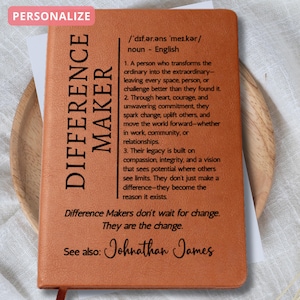 May include: A brown leather-bound notebook with the words "DIFFERENCE MAKER" in large, bold letters. The notebook includes a definition and inspirational text. The word "PERSONALIZE" is in a pink box in the upper left corner.
