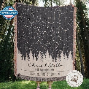 May include: A dark gray woven blanket featuring a constellation design with white stars and lines. The bottom of the blanket has a forest silhouette and the text "Chris & Stella, OUR WEDDING DAY, AUGUST 12, 2023 I ST. LOUIS, MO". The blanket has fringed edges.