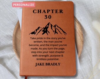 以下が含まれることがあります： 「CHAPTER 30」の文字と山脈のグラフィックが描かれた茶色の革製ジャーナル。グラフィックの下のテキストには、「Take pride in the story you've written, the man you've become, and the impact you've made. As you turn the page, step into your next chapter with strength, purpose & limitless potential.」と書いてあります。一番下のテキストには「JAKE BRADLY」と書いてあります。