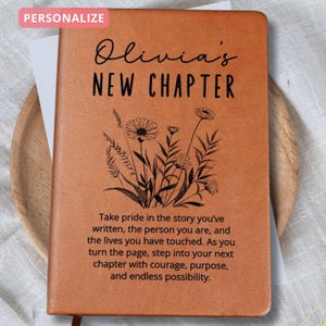 Könnte beinhalten: Ein personalisiertes braunes Lederjournal mit dem Text "Olivia's NEW CHAPTER" und einem floralen Design. Das Journal enthält inspirierenden Text. Das Wort "PERSONALIZE" befindet sich in einer rosa Box oben links.