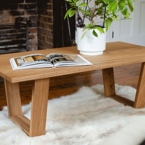 May include: A rectangular wooden coffee table with a light brown finish. The table has a modern design with angled legs and a smooth surface. An open book and a potted plant sit on top of the table, which is placed on a white fur rug.