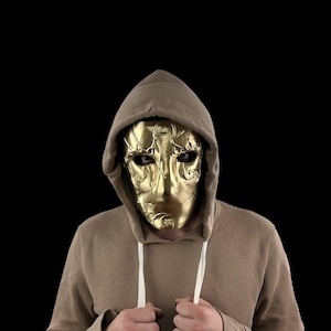 May include: A person wearing a brown hooded sweatshirt with a gold mask covering their face. The mask has intricate details and eye holes.