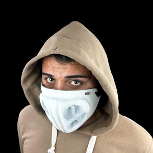 Peut inclure: Un masque facial blanc au design unique, porté avec un sweat à capuche beige. Le masque présente un aspect sculpté avec de multiples ouvertures. Le fond est noir.
