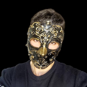 May include: A black masquerade mask with gold accents and intricate details. The mask covers the entire face, with eye holes and a small nose opening.