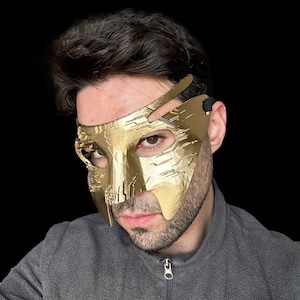 Op de afbeelding: Een gouden masker met een geometrisch ontwerp. Het masker bedekt de ogen en een deel van de neus en heeft een band die om het hoofd gaat.