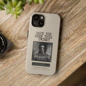Puede incluir: Funda de teléfono beige con el texto "HAVE YOU SEEN THIS TWINK?" y una imagen en blanco y negro de una persona sosteniendo un letrero de prisión. Una lente de cámara negra y una planta verde son visibles.