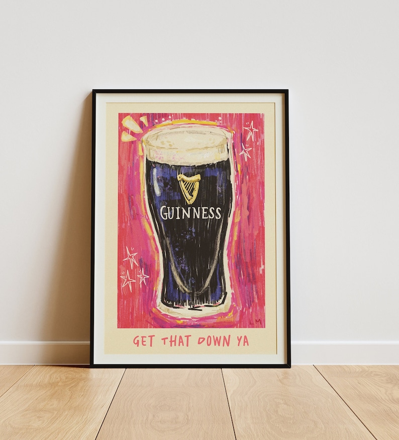 Puede incluir: Un p&oacute;ster enmarcado con fondo rosa y una ilustraci&oacute;n de dibujos animados de un vaso de cerveza Guinness. El texto "GET THAT DOWN YA" est&aacute; en la parte inferior del p&oacute;ster.