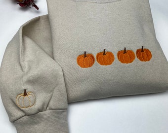 Könnte beinhalten: Ein hellgrauer Pullover mit einer Reihe von vier gestickten orangefarbenen Kürbissen auf der Vorderseite. Der Pullover hat lange Ärmel und einen Rundhalsausschnitt.