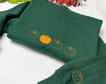 May include: A dark green sweatshirt with a row of five embroidered pumpkins in orange and brown. The pumpkins are arranged in a line across the front of the sweatshirt.