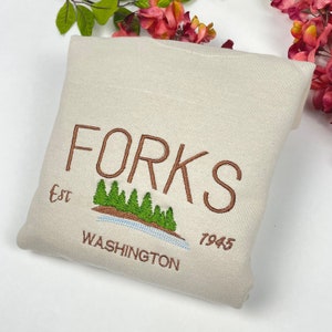 May include: A beige sweatshirt with brown embroidered text that reads "FORKS Est. 7945 WASHINGTON" with a small embroidered image of a lake and trees.