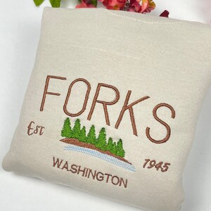 May include: A beige sweatshirt with brown embroidery that reads "FORKS Est. 7945 WASHINGTON" with a green tree graphic.