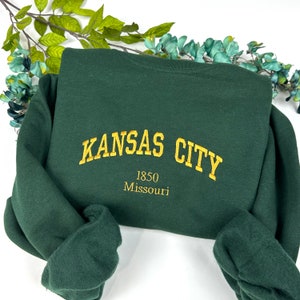 Op de afbeelding: Een donkergroen sweatshirt met gouden geborduurde tekst die "KANSAS CITY 1850 Missouri" luidt.
