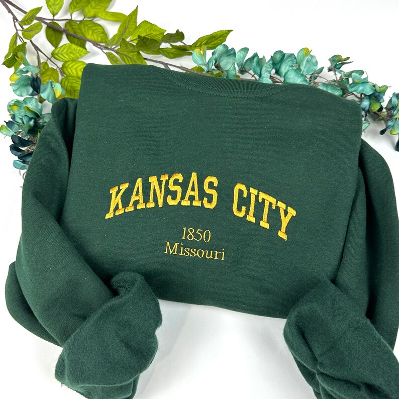 Op de afbeelding: Een donkergroen sweatshirt met gouden geborduurde tekst die "KANSAS CITY 1850 Missouri" luidt.