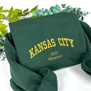 Op de afbeelding: Een donkergroen sweatshirt met de tekst "KANSAS CITY 1850 Missouri" geborduurd in goud op de achterkant.