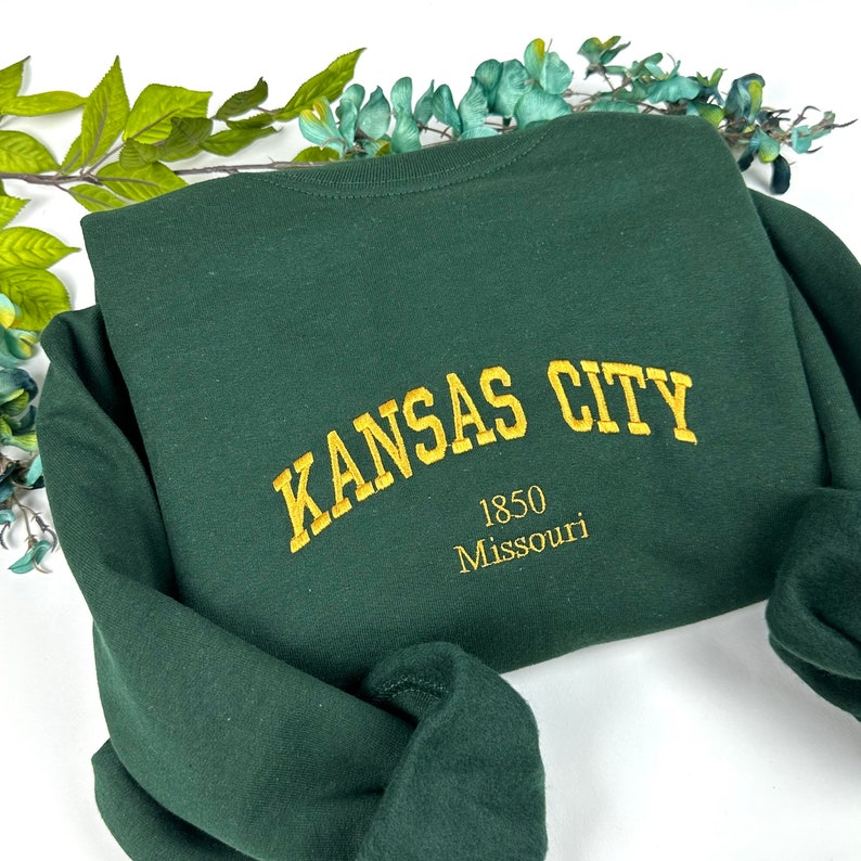 Op de afbeelding: Een donkergroen sweatshirt met de tekst "KANSAS CITY 1850 Missouri" geborduurd in goud op de achterkant.