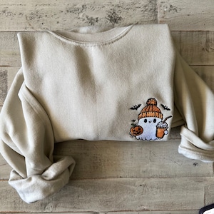 May include: A light beige sweatshirt with an embroidered design of a ghost wearing a brown and orange hat, holding a pumpkin and a cup of coffee. The ghost is surrounded by bats.