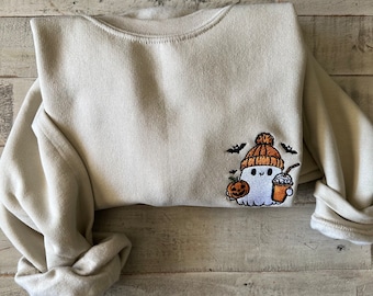 May include: A light beige sweatshirt with an embroidered design of a ghost wearing a brown and orange hat, holding a pumpkin and a cup of coffee. The ghost is surrounded by bats.