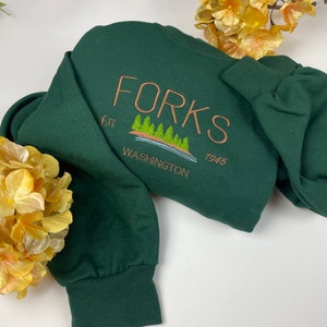 May include: A dark green sweatshirt with the text "FORKS est 1945 WASHINGTON" embroidered in brown on the back. The embroidery also includes a small image of a lake with trees.
