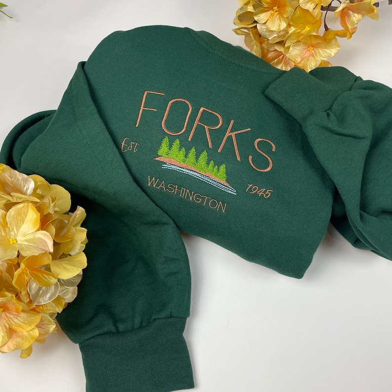 May include: A dark green sweatshirt with the text "FORKS est 1945 WASHINGTON" embroidered in brown on the back. The embroidery also includes a small image of a lake with trees.